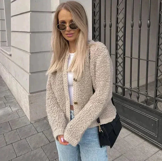 Frau in beiger Teddyjacke, weißem Top, blauer Jeans, Sonnenbrille, schwarze Tasche, draußen.
