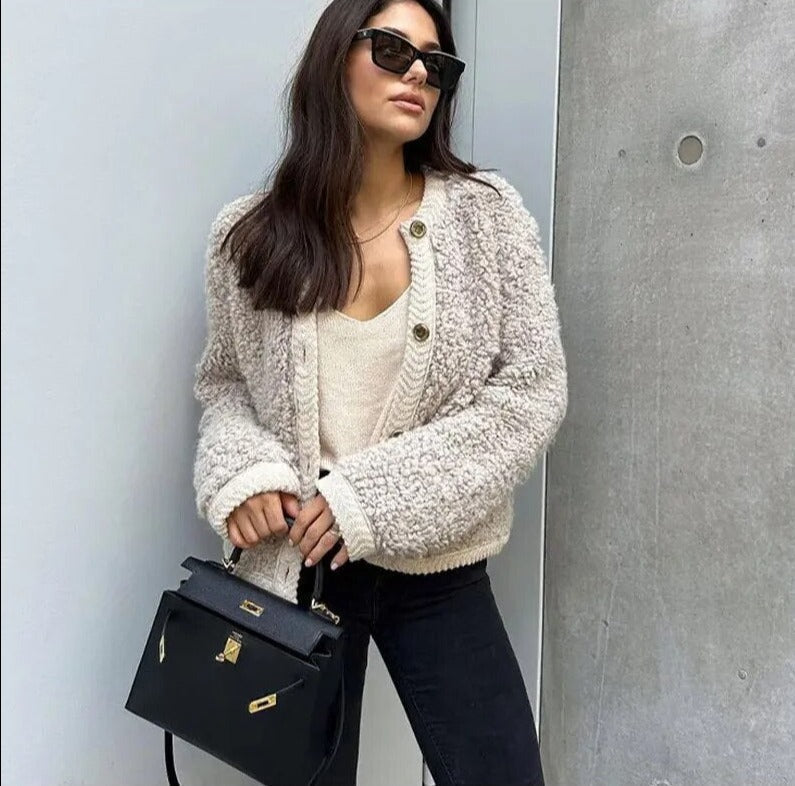 Frau mit beiger Strickjacke, schwarzer Handtasche und Sonnenbrille vor grauer Wand.