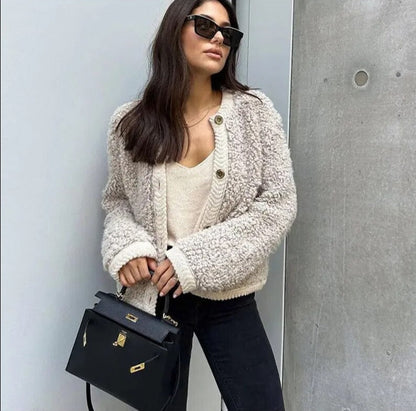 Frau mit beiger Strickjacke, schwarzer Handtasche und Sonnenbrille vor grauer Wand.