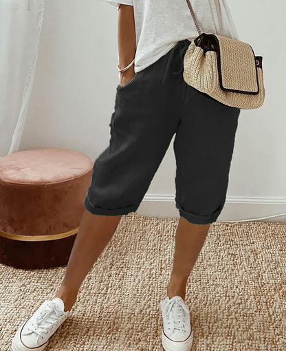 Frau in schwarzer Bermuda-Shorts, weißem T-Shirt, Strohtasche, weiße Sneaker, lässiger Stil.
