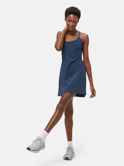 Frau in dunkelblauem Sportkleid, lächelnd, mit grauen Laufschuhen, auf weißem Hintergrund.