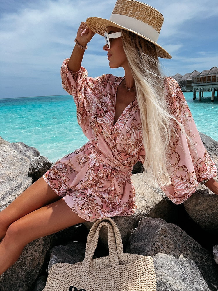 Frau in rosa Sommerkleid und Strohhut am Strand, sitzt auf Felsen vor türkisfarbenem Meer.