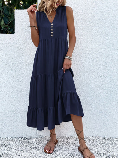 Frau in dunkelblauem Sommerkleid mit Knöpfen, ärmellos, knielang, Sandalen, vor weißer Wand.