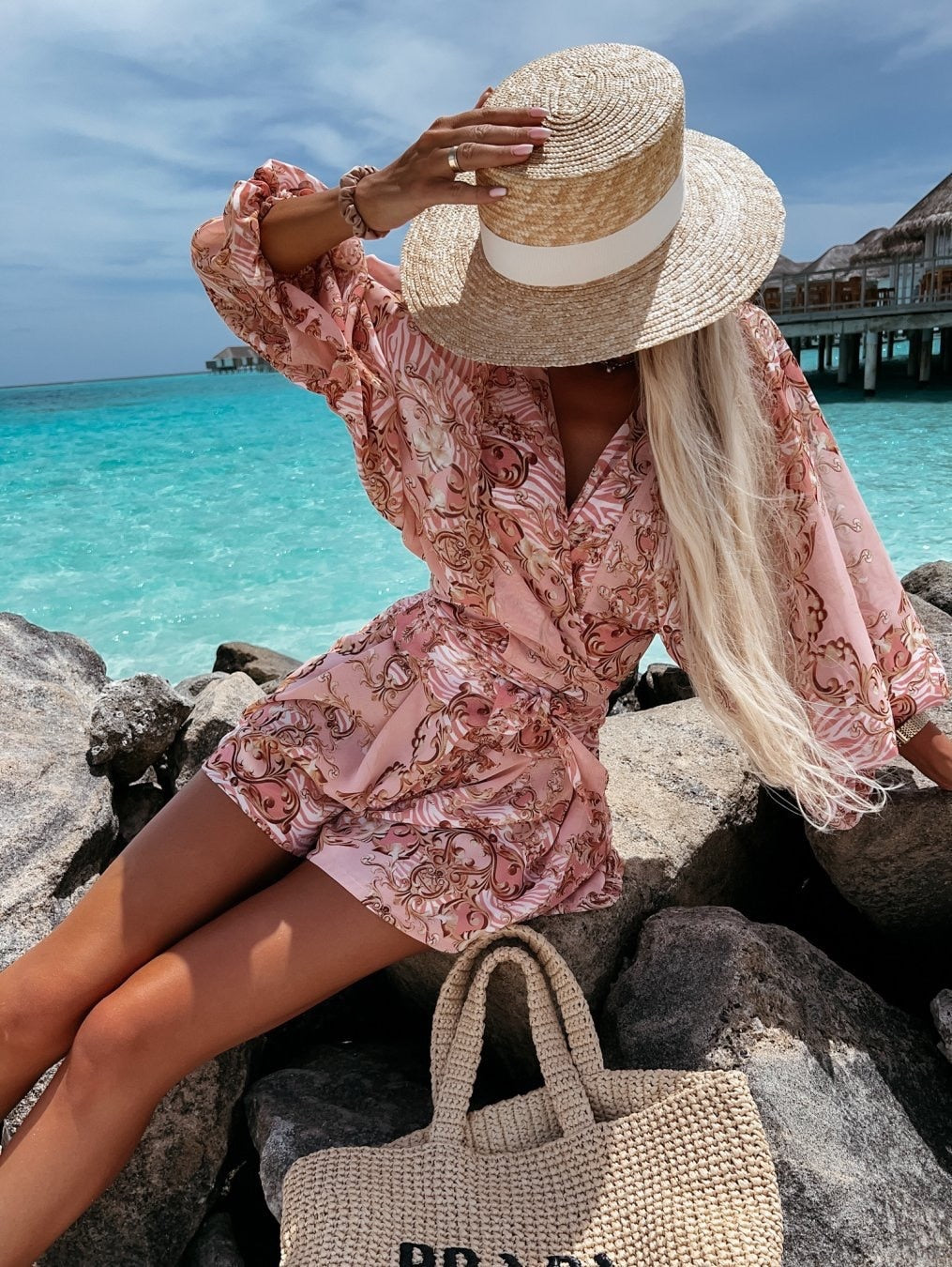 Frau in rosa Sommerkleid mit Strohhut am Strand, türkisfarbenes Meer im Hintergrund.