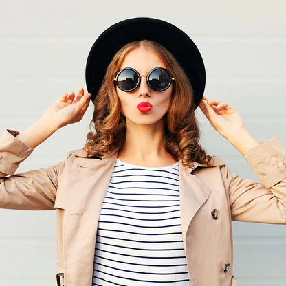 Frau mit schwarzem Hut, runder Sonnenbrille, gestreiftem Shirt und beigem Trenchcoat.
