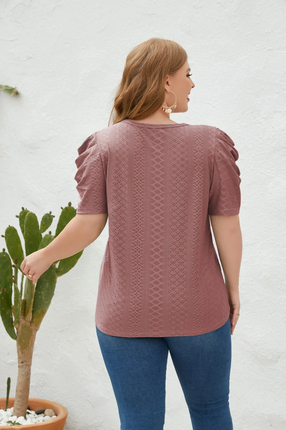 Frau in rosa Spitzenbluse mit Puffärmeln, blaue Jeans, vor weißer Wand, neben Kaktus.