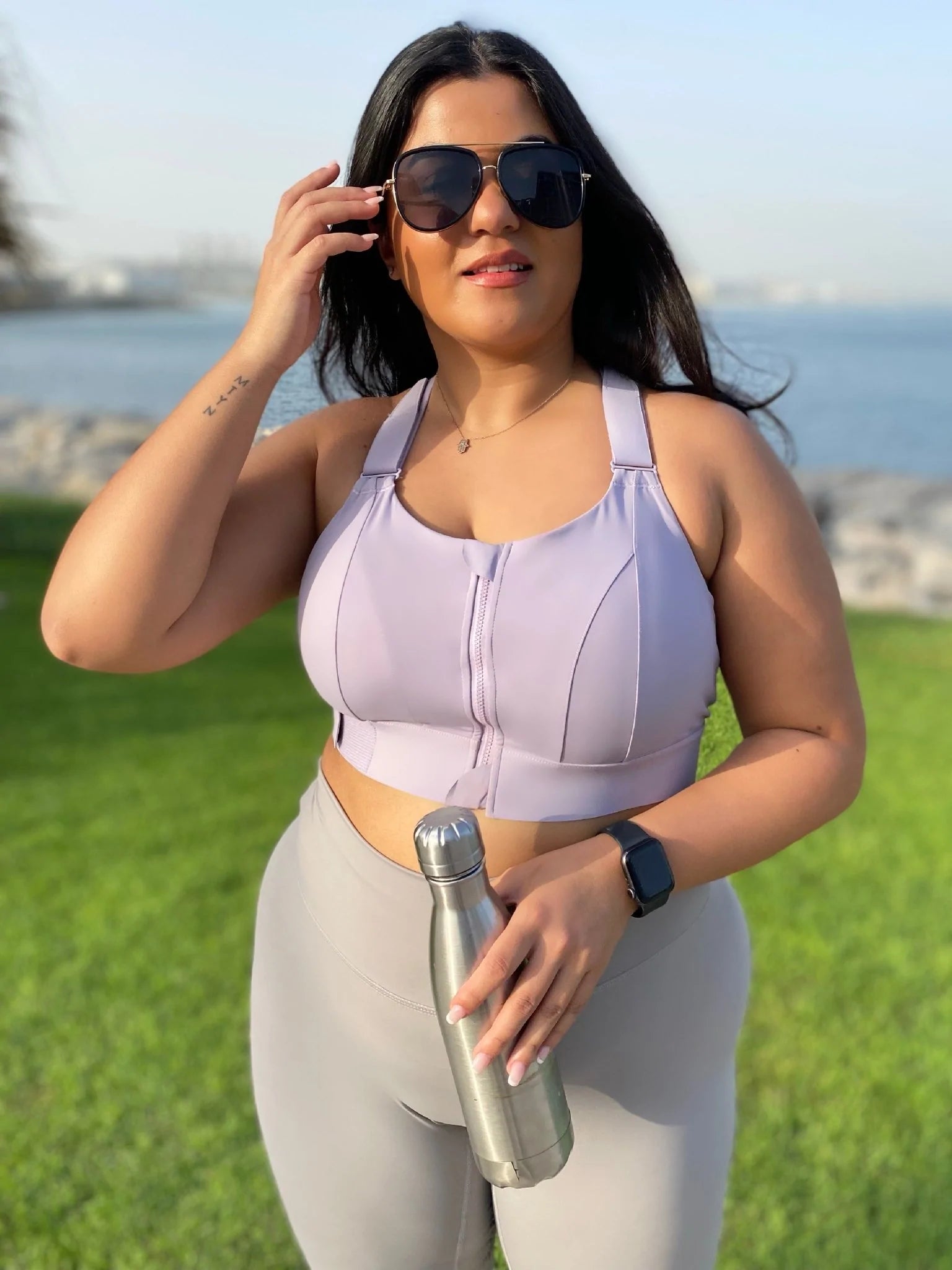 Frau in lila Sport-BH und grauer Leggings mit Wasserflasche im Freien, Fitnessmode.