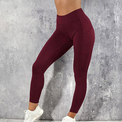 Frau in bordeauxroter Sportleggings, hohe Taille, vor grauer Wand, weiße Turnschuhe.