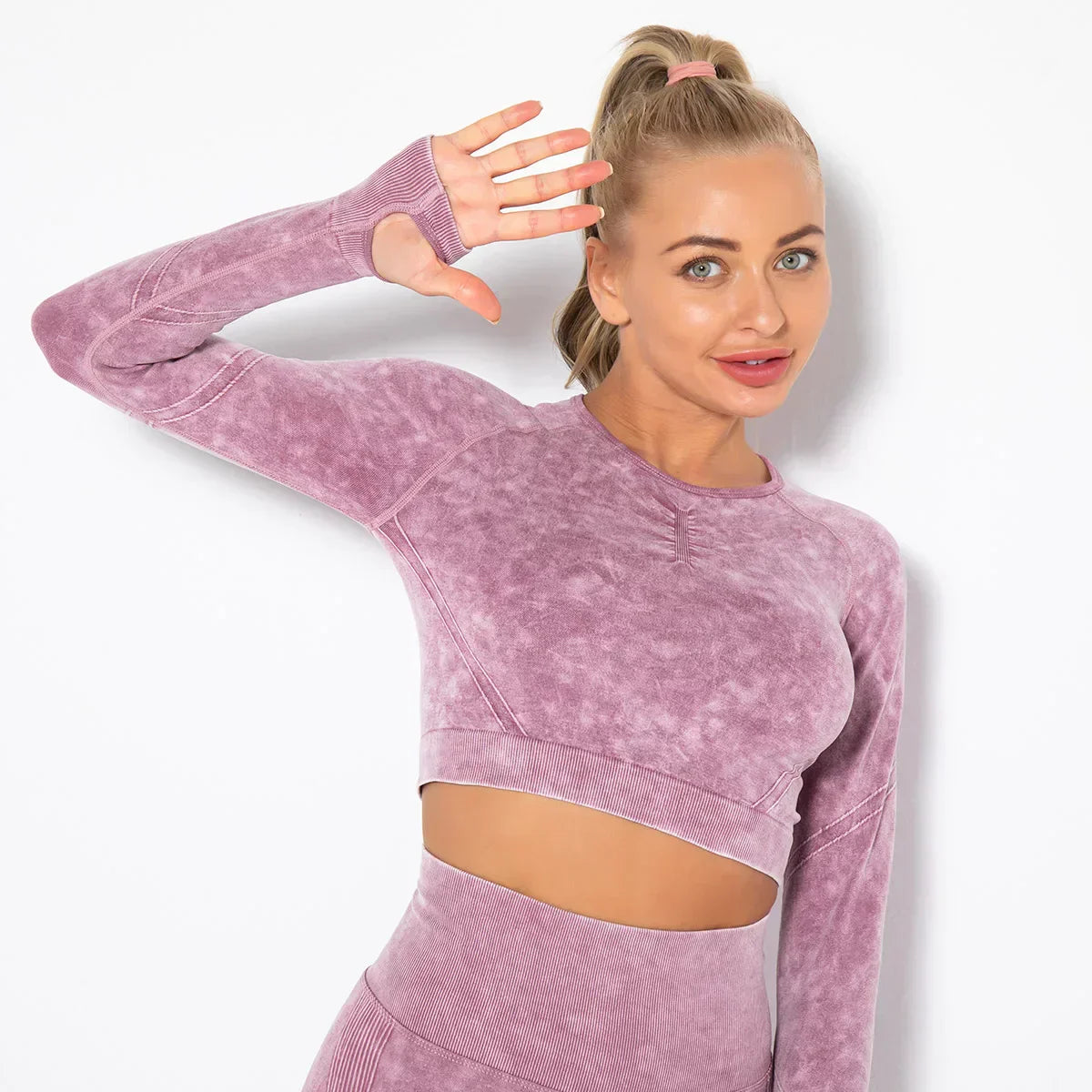 Frau in lila Sport-Set, nahtloses Langarm-Oberteil, Fitnessmode, lächelnd, Hand erhoben.