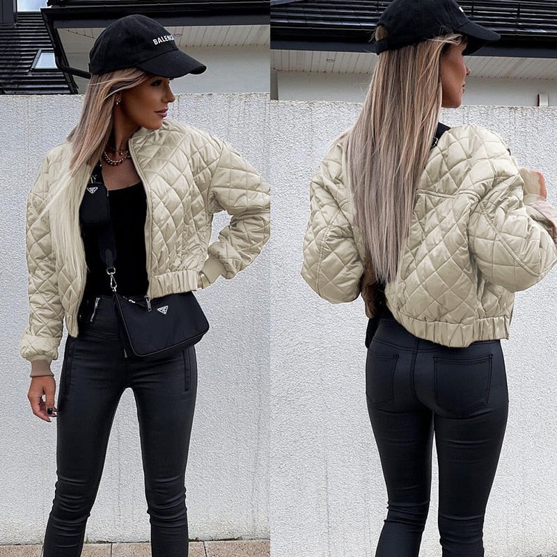 Frau in beiger Steppjacke, schwarzer Hose, schwarzem Top und schwarzer Kappe vor weißer Wand.
