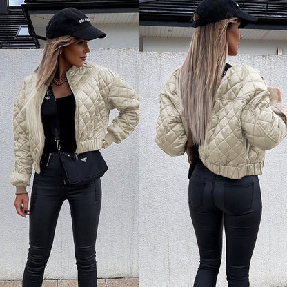 Frau in beiger Steppjacke, schwarzer Hose, schwarzem Top und schwarzer Kappe vor weißer Wand.
