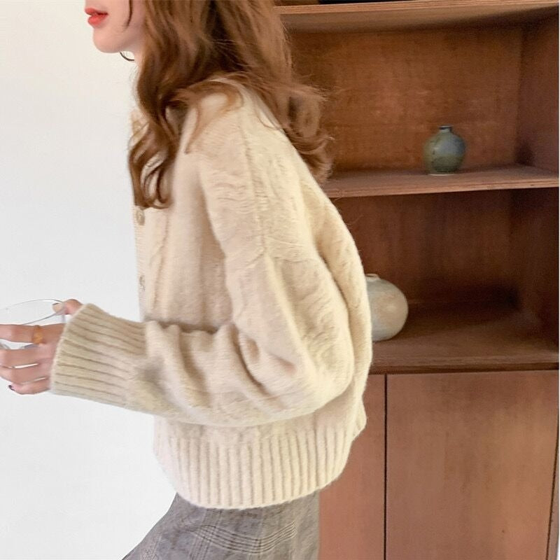 Frau in beigem Strickpullover, hält Glas, vor Holzregal mit Keramikvasen.