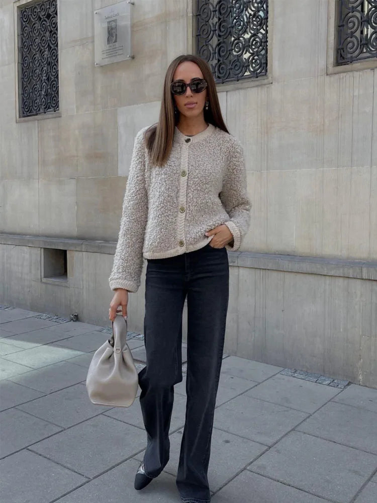 Frau in beiger Strickjacke, schwarzer Hose, Sonnenbrille, mit grauer Handtasche auf Gehweg.