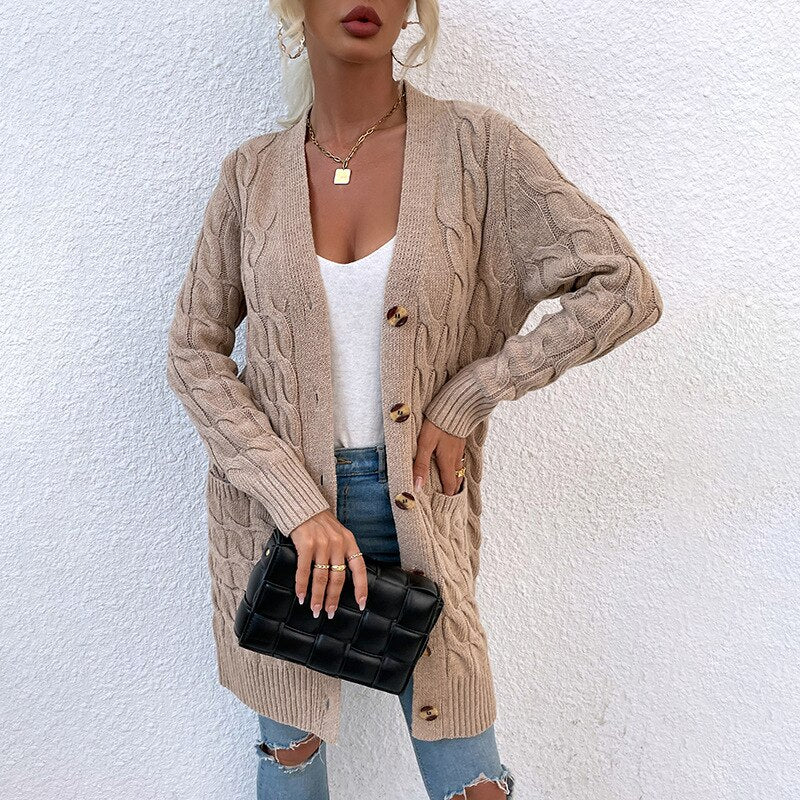 Frau in beigem Strickcardigan mit Zopfmuster, weiße Bluse, zerrissene Jeans, schwarze Clutch.