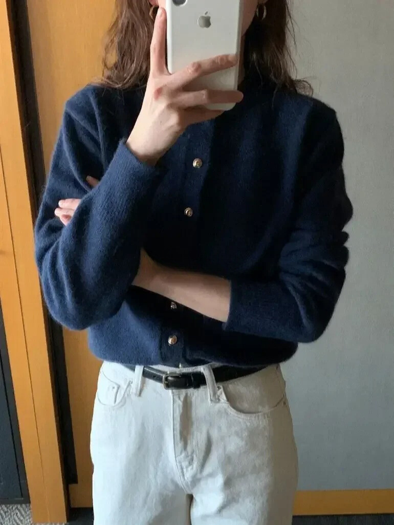 Frau in dunkelblauem Pullover und weißer Hose macht Selfie mit Smartphone.