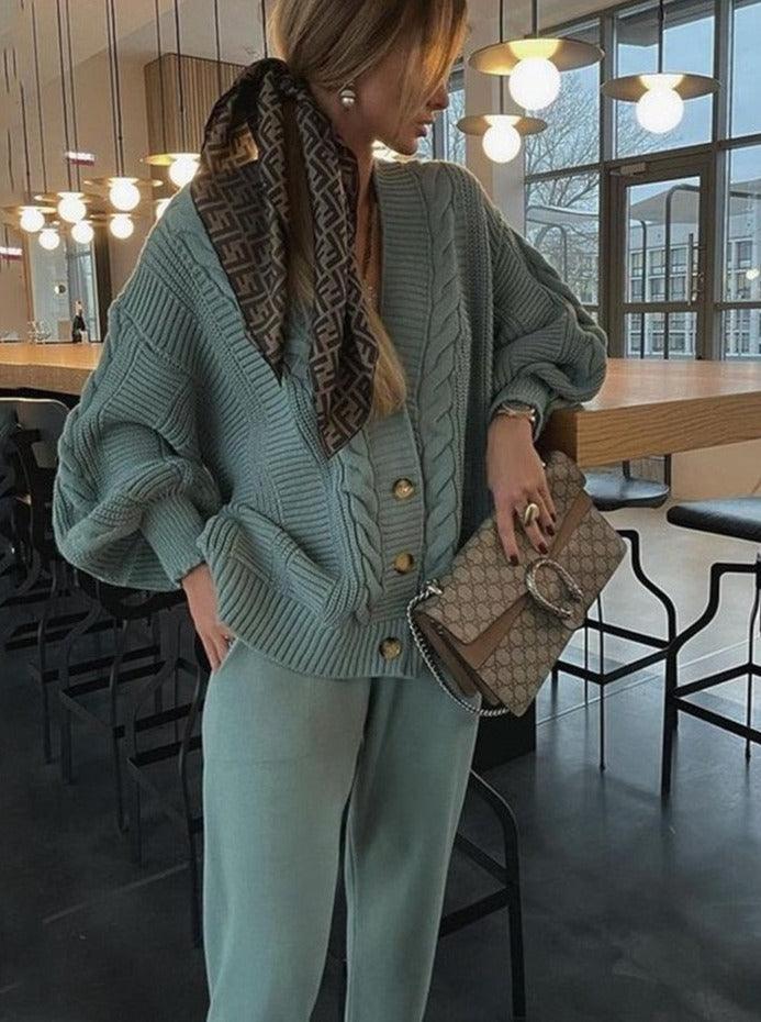 Frau in grünem Strickpullover und Hose, hält Designer-Handtasche, modisches Outfit, Innenraum.