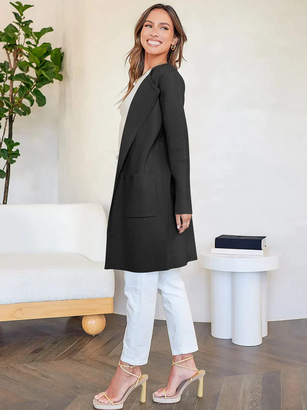 Frau in schwarzem Cardigan, weißer Hose, beiger High Heels, lächelnd, moderne Wohnzimmerkulisse.