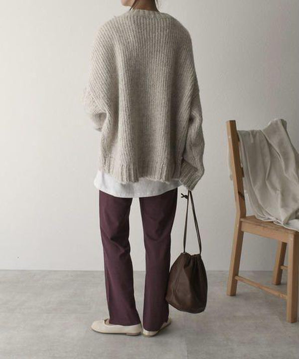 Frau in beigem Strickpullover, lila Hose, braune Tasche, steht neben Holzstuhl.