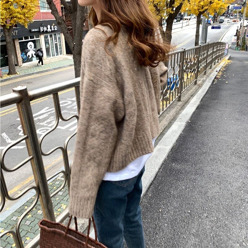 Frau in braunem Strickpullover auf Gehweg im Herbst, trägt braune Tasche.