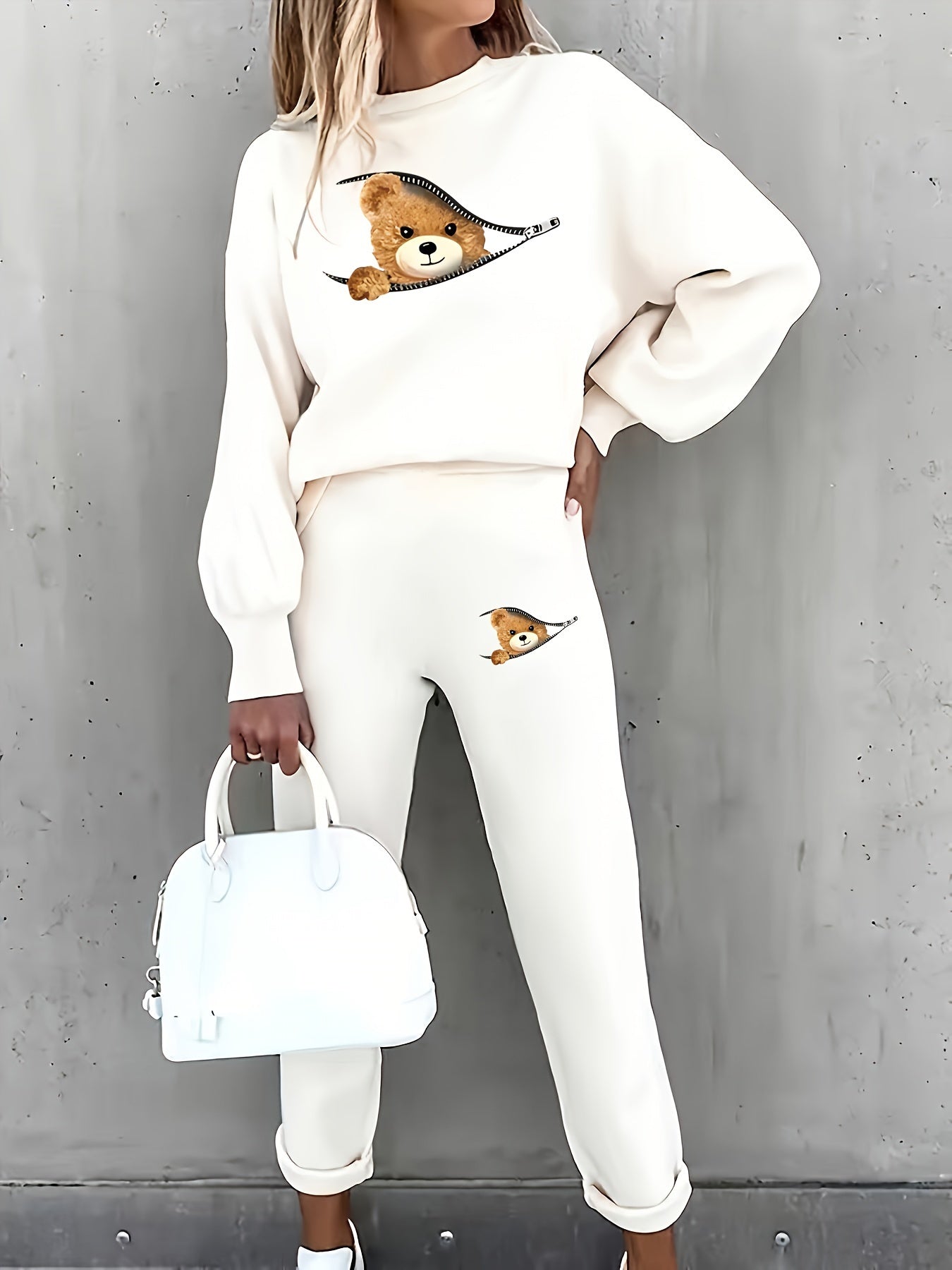 Frau in cremefarbenem Teddy-Print-Sweatshirt und Jogginghose, hält weiße Handtasche.