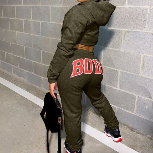 Frau in olivgrünem Jogginganzug mit "BODY"-Aufdruck, Kapuze, Turnschuhe, Betonwand.