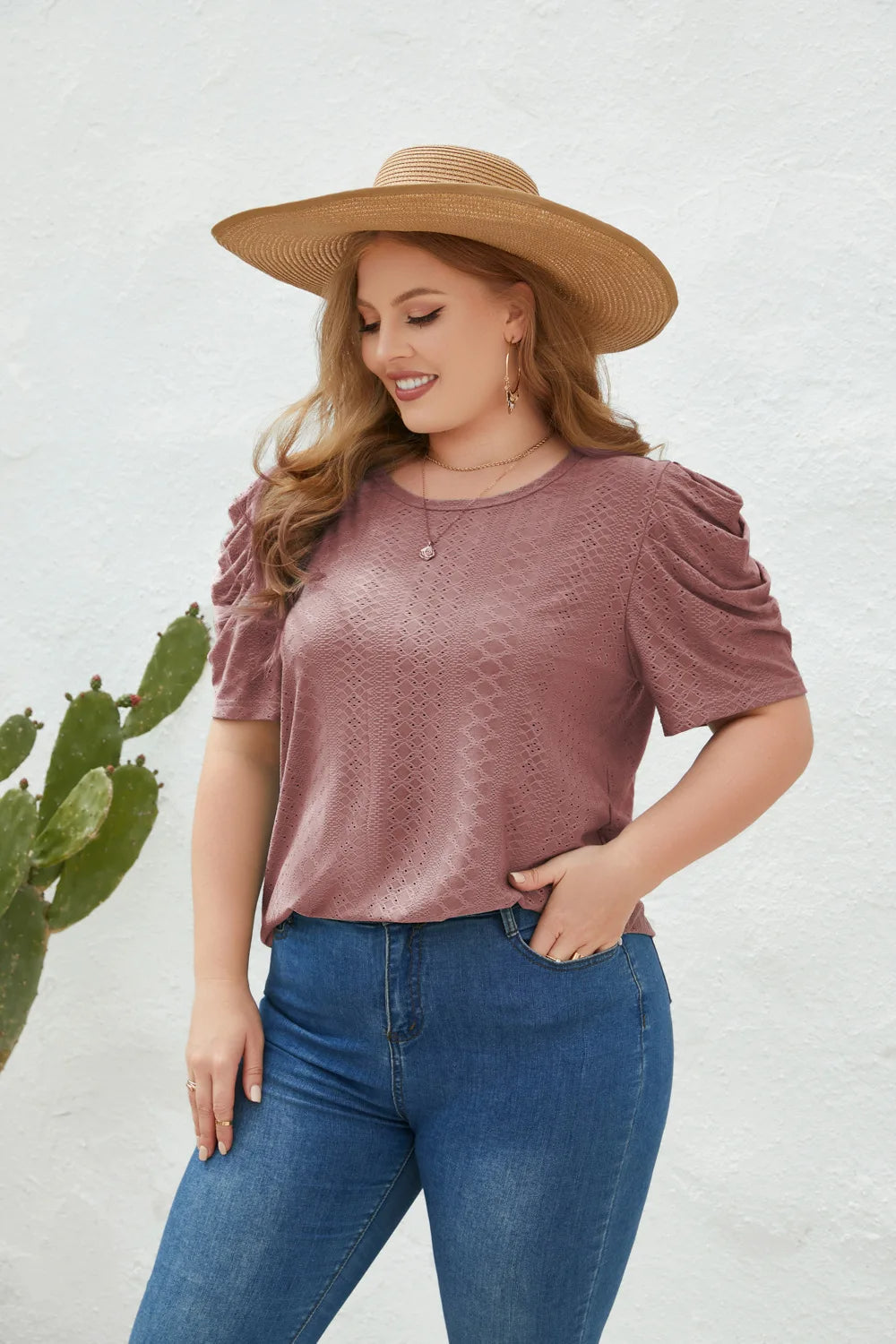 Frau in rosa Spitzenbluse und Strohhut, lächelnd, vor weißer Wand und Kaktus.