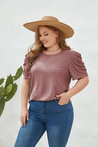 Frau in rosa Spitzenbluse und Strohhut, lächelnd, vor weißer Wand und Kaktus.