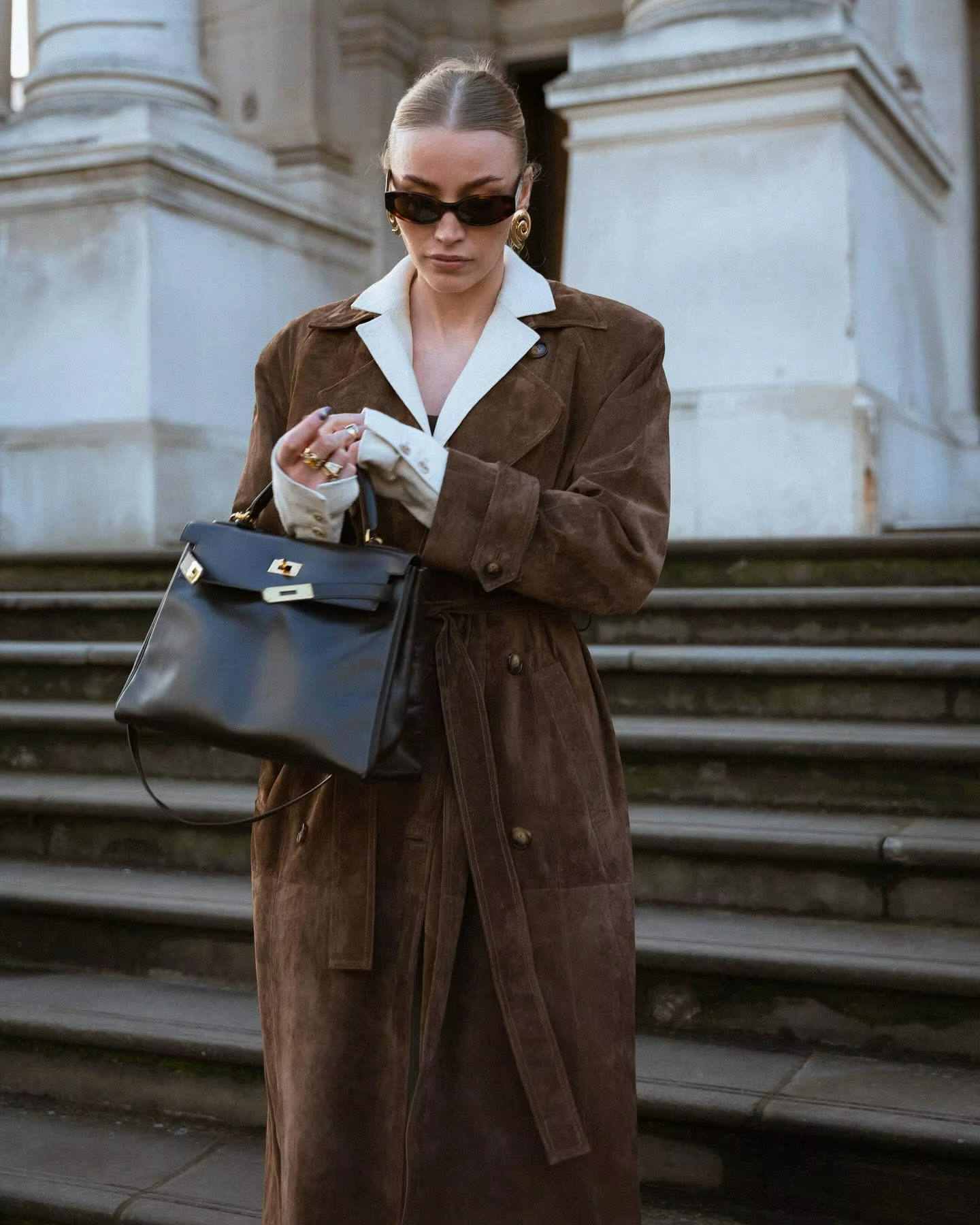 Frau in braunem Wildledermantel mit schwarzer Ledertasche und Sonnenbrille auf Treppe.