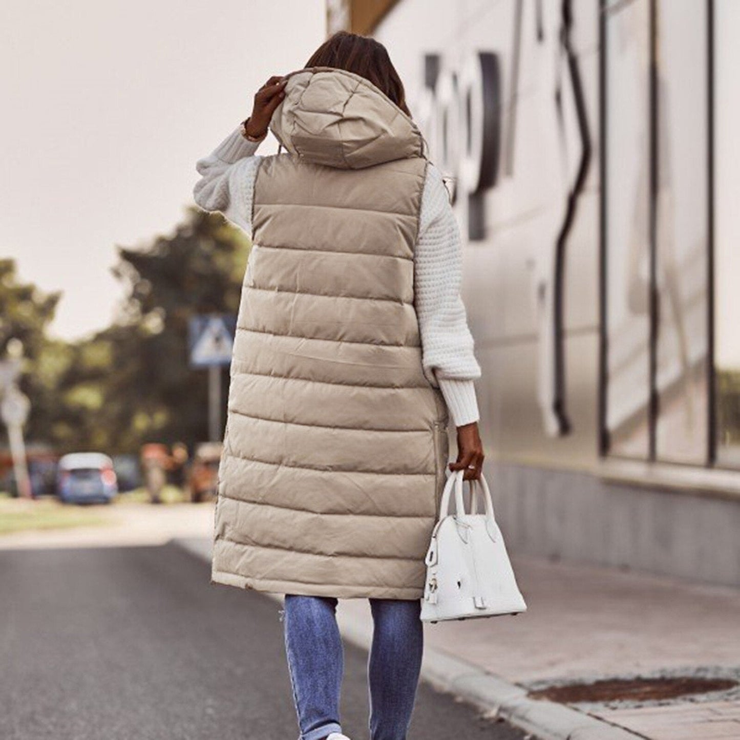 Frau in beiger Steppweste mit Kapuze, weiße Handtasche, lässiger Herbstlook, Straße im Hintergrund.
