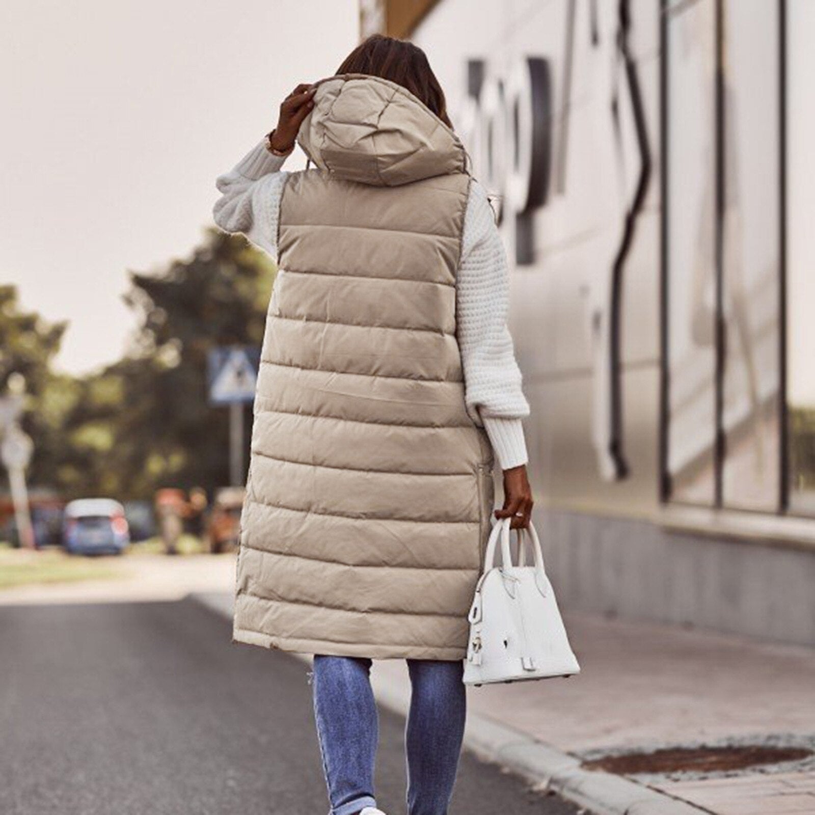 Frau in beiger Steppweste mit Kapuze, weiße Handtasche, lässiger Herbstlook, Straße im Hintergrund.