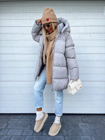Frau in grauer Daunenjacke, Jeans, Mütze und Stiefeln vor Holzwand.