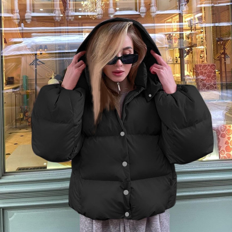 Frau in schwarzer Daunenjacke mit Kapuze und Sonnenbrille vor Schaufenster.