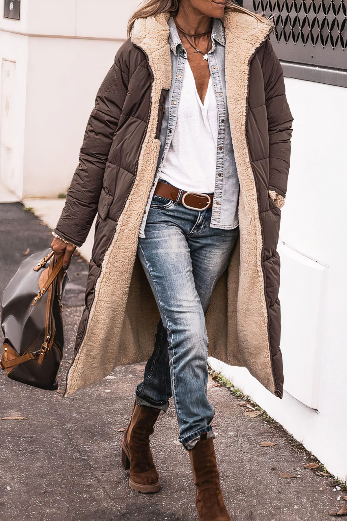 Frau in braunem Daunenmantel mit Lammfellfutter, Jeans und Stiefeln, trägt braune Tasche.