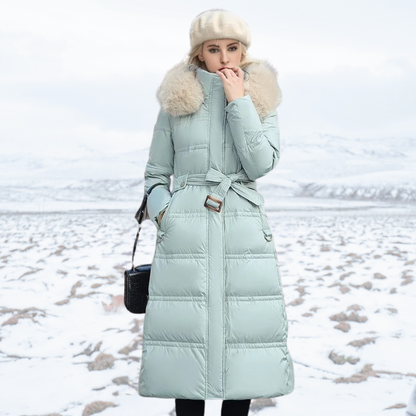 Frau in mintgrünem Daunenmantel mit Kapuze und Gürtel im verschneiten Winterlandschaft.