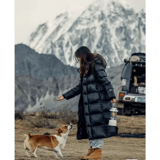 Frau im langen schwarzen Daunenmantel mit Hund vor schneebedecktem Berg und Geländewagen.