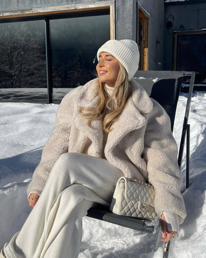Frau in weißem Wintermantel und Mütze, sitzt im Schnee, hält Designer-Handtasche.