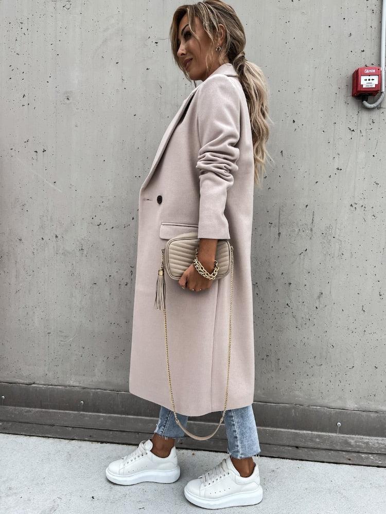 Frau in beigem Wollmantel, Jeans, weißen Sneakers, mit beiger Tasche, vor Betonwand.