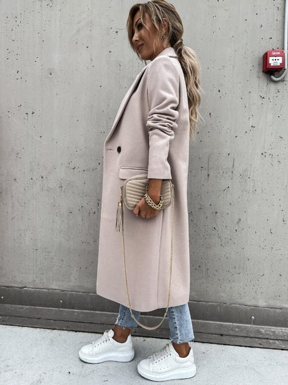 Frau in beigem Wollmantel, Jeans, weißen Sneakers, mit beiger Tasche, vor Betonwand.