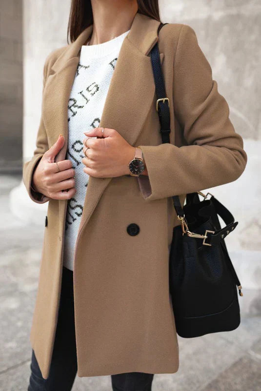 Dame in beigem Mantel, weißem Pullover, schwarzer Tasche und Uhr, modisch elegant gekleidet.