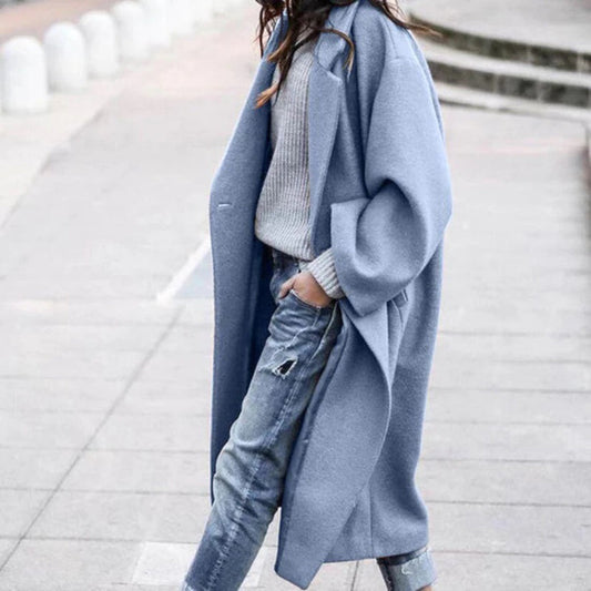 Frau in blauem Wollmantel, grauem Pullover und Jeans auf Gehweg spazierend.