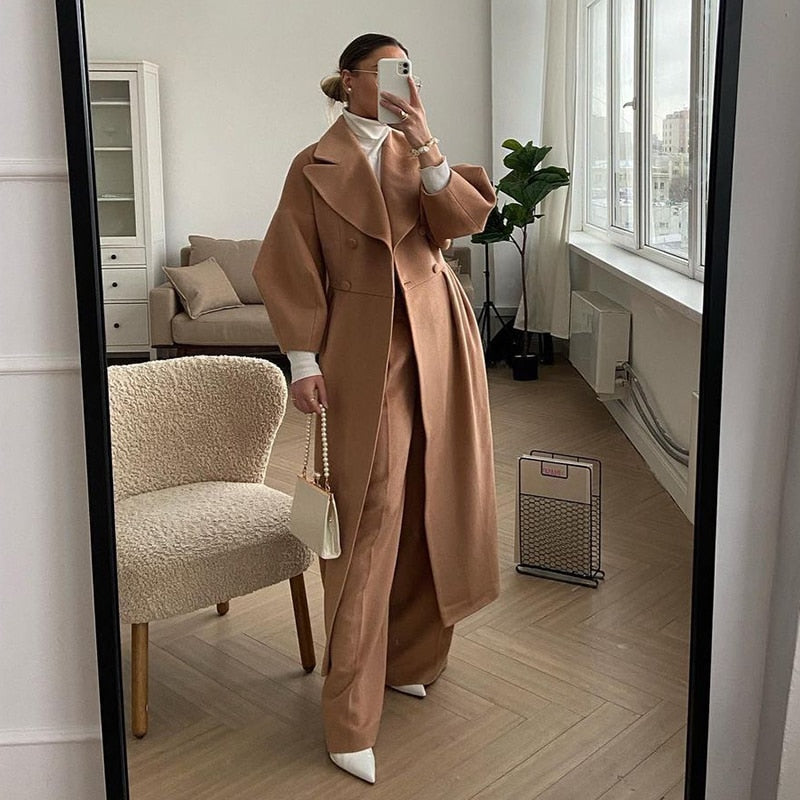 Frau im eleganten, beigen Wollmantel, weiße Stiefel, Handtasche, modernes Wohnzimmer.