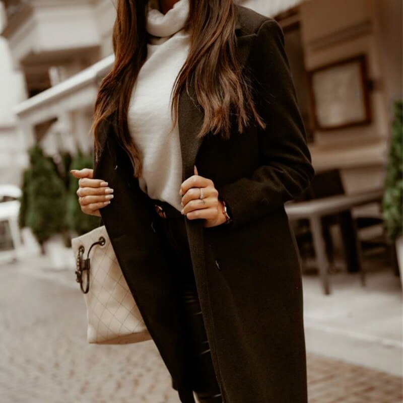 Frau in schwarzem Mantel und weißem Rollkragenpullover, hält beige Handtasche, Stadtstraße.