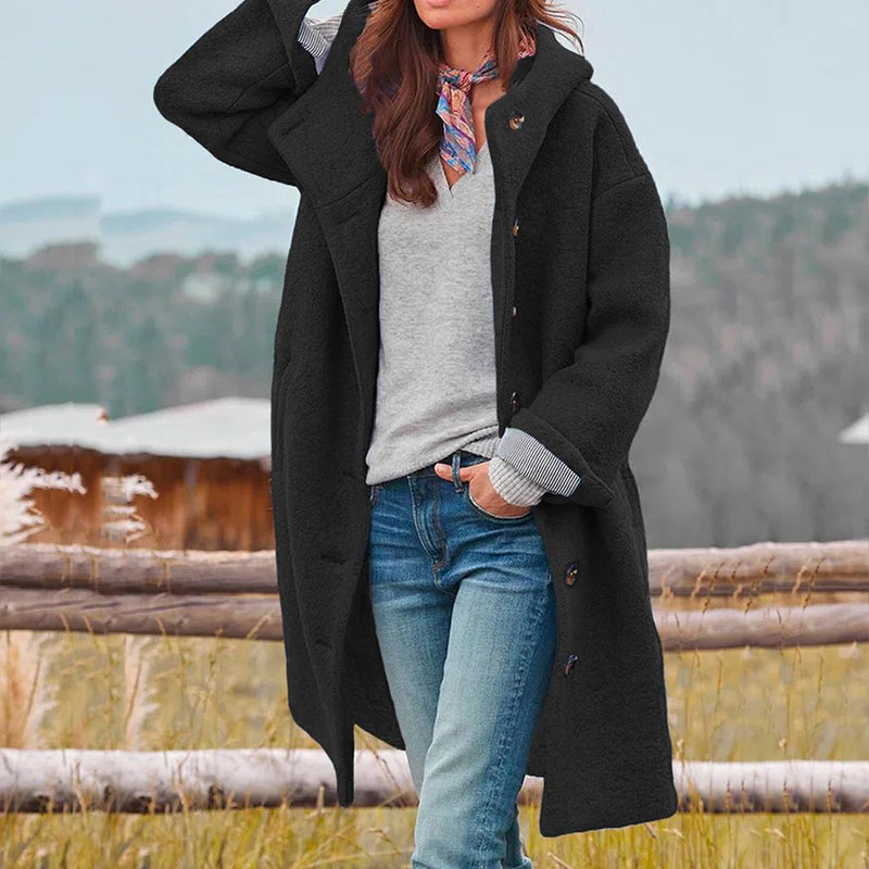 Frau in schwarzem Wollmantel, grauem Pullover und Jeans vor ländlicher Kulisse.