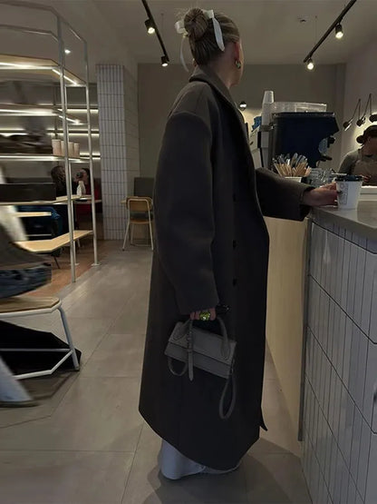 Frau im Café mit langem, dunkelgrauem Mantel und kleiner Handtasche, moderne Inneneinrichtung.
