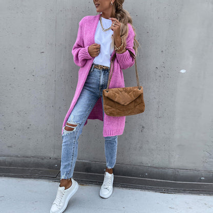 Frau in rosa Strickjacke, Jeans und weißen Sneakers mit brauner Umhängetasche vor grauer Wand.