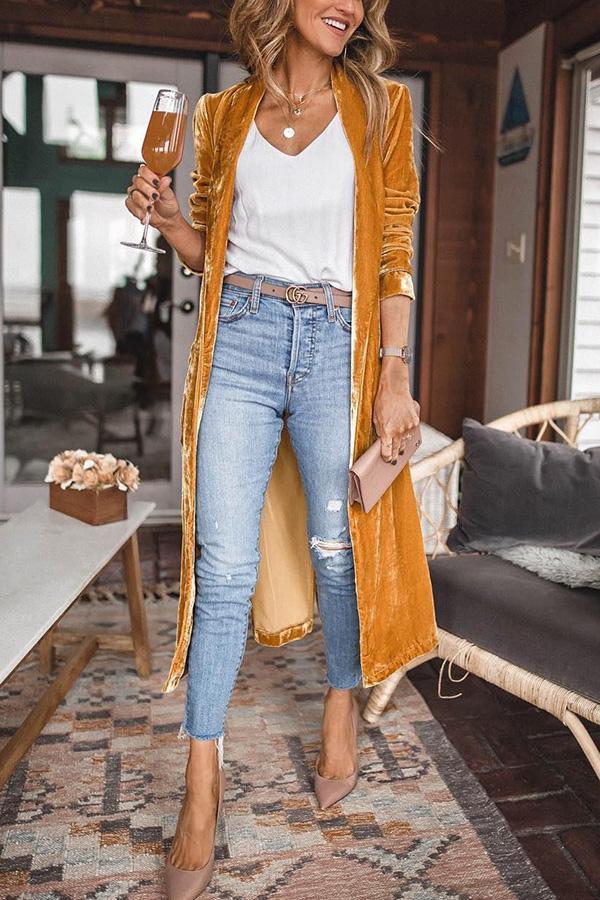 Frau in senfgelbem Samt-Cardigan, Jeans und Pumps, hält ein Glas, modisch, lässiger Stil.