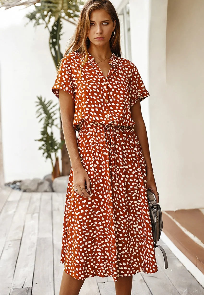 Frau in rotem Sommerkleid mit weißen Punkten, stehend auf Holzterrasse, modisch, elegant.