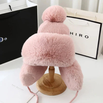 Rosa Wintermütze mit Kunstfell, Bommel und Ohrenklappen auf Holzständer, modisch und warm.