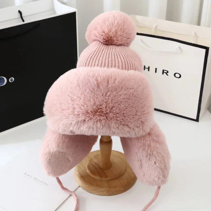 Rosa Wintermütze mit Kunstfell, Bommel und Ohrenklappen auf Holzständer, modisch und warm.
