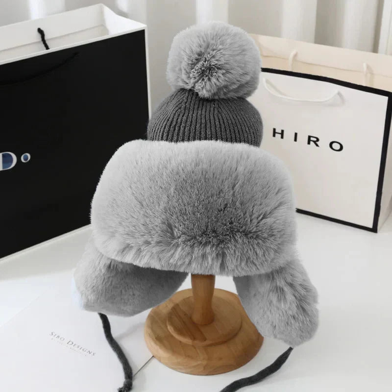 Graue Wintermütze mit Kunstfell, Bommel und Ohrenklappen auf Holzständer, warm und modisch.